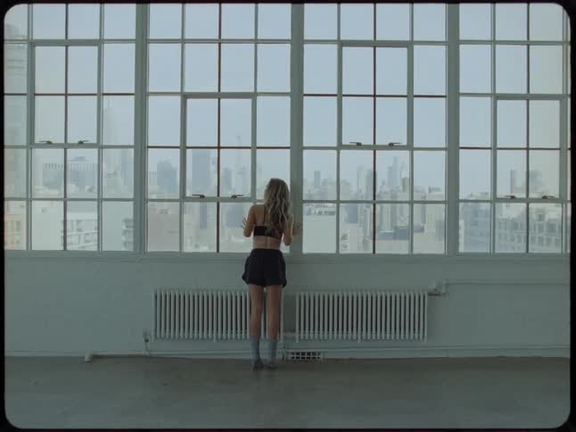 A solitary woman gazes out of a large industrial window at the city skyline from an empty loft apartment