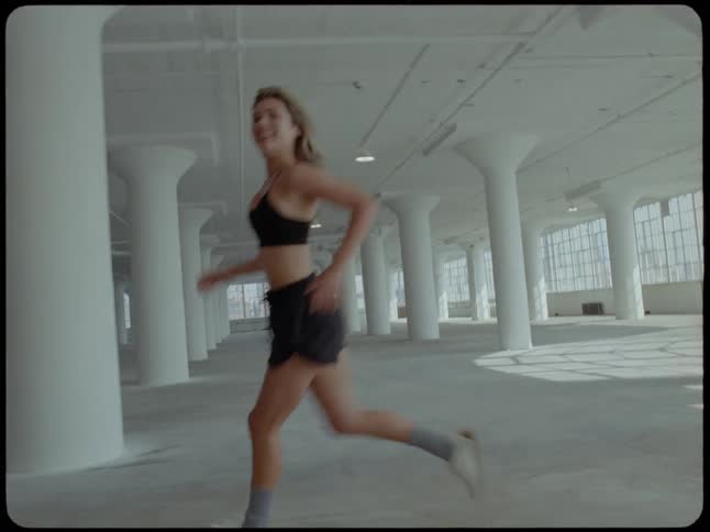 A smiling woman in sportswear runs in slow motion through a vast, sunlit industrial loft with white pillars