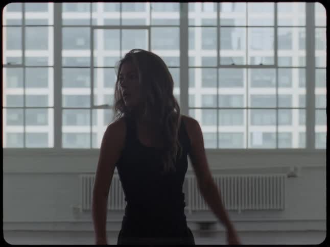 A silhouetted woman walks slowly and gracefully through an empty industrial loft with large windows