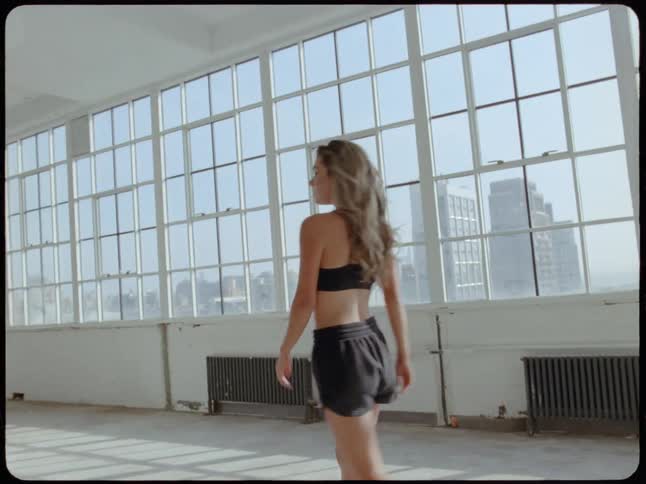 A graceful woman in athletic wear walks in slow motion through a sun-drenched industrial loft with city views
