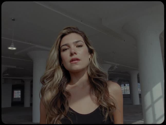A beautiful woman tilts her head back to feel the sunlight in a warehouse before smiling at the camera