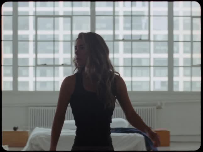 Super 8mm style footage shows a woman in a black tank top walking through her bright city loft apartment