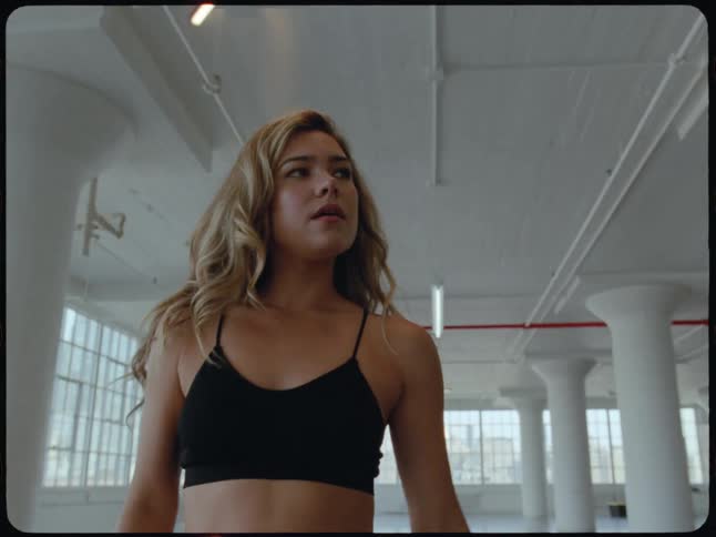 Low-angle slow motion shot of a focused female dancer letting her hair down and staring intensely at the camera in a large industrial studio