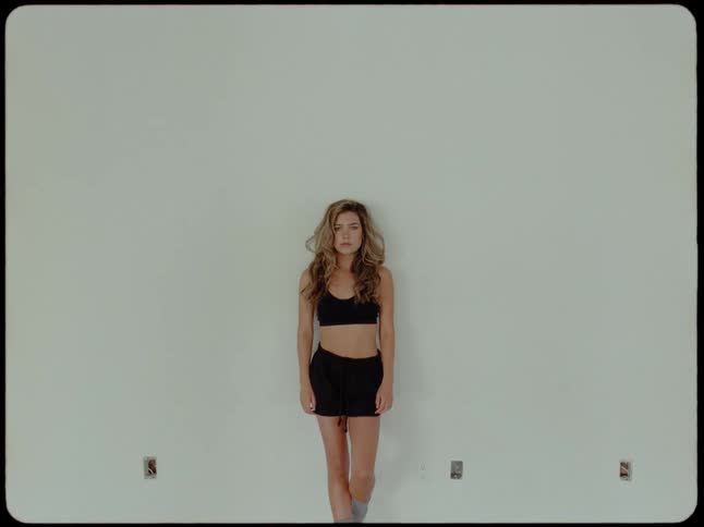 A vintage film-style shot of a young woman in athletic wear standing against a blank wall, slowly turning to look seriously into the camera