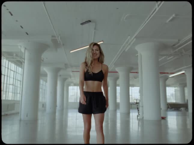 A confident woman in athletic wear walks and playfully twirls in a spacious, sunlit industrial loft studio