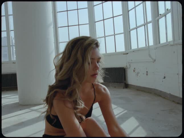 A graceful woman flips her long, flowing hair in slow motion while sitting thoughtfully in a sunlit industrial loft