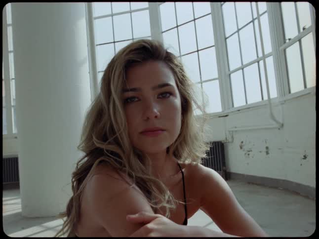 A beautiful young woman with long wavy hair sits on the floor of a sunlit industrial loft and turns to look into the camera