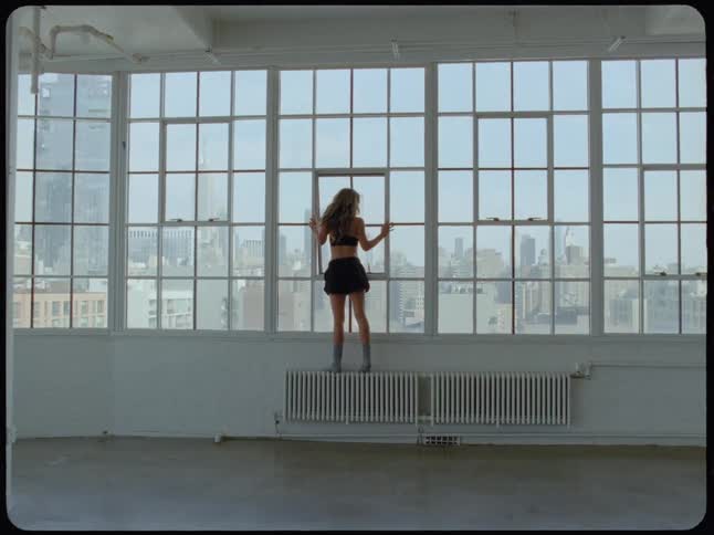 A woman stands on a radiator in a minimalist loft, gazing out at the vast city skyline through a wall of windows