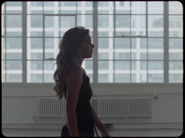 A thoughtful young woman walks in slow motion through a spacious urban loft, her silhouette framed by large, bright windows
