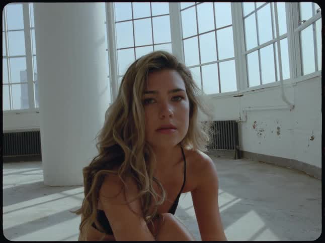A beautiful woman with long wavy hair sits on the floor of a sun-drenched loft and gazes thoughtfully at the camera