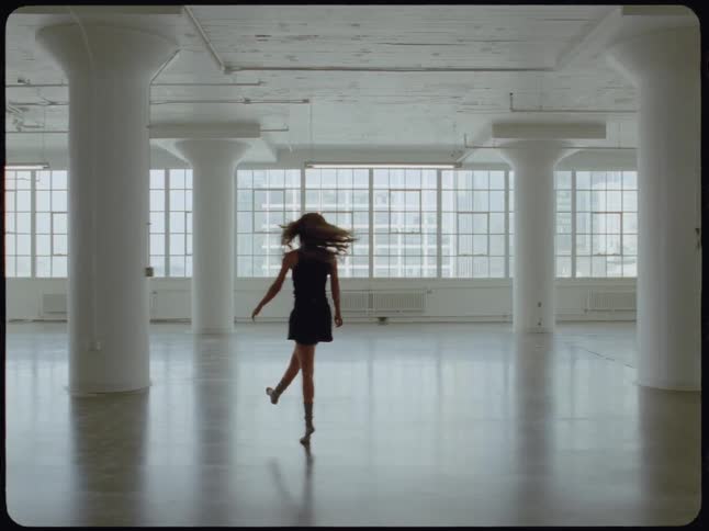 A solitary woman walks and dances expressively in a vast, empty white studio with large windows