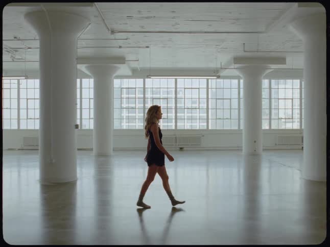 A woman in a black outfit walks alone through a large, empty, naturally lit urban loft space