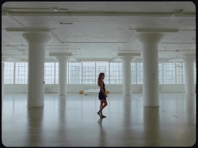 A solitary woman in a black dress walks through a spacious, empty white industrial loft, her figure moving between large pillars