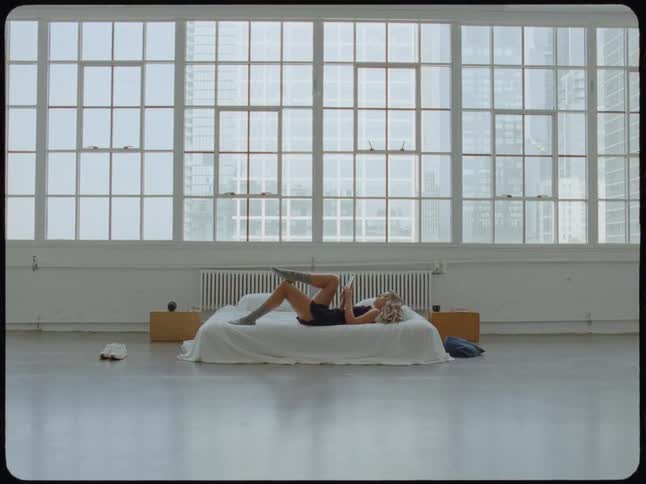 A woman relaxes on a bed reading a book in a spacious, minimalist loft apartment with large windows overlooking a city