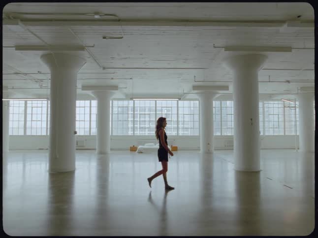 A solitary woman walks barefoot across the polished floor of a vast, empty white industrial loft