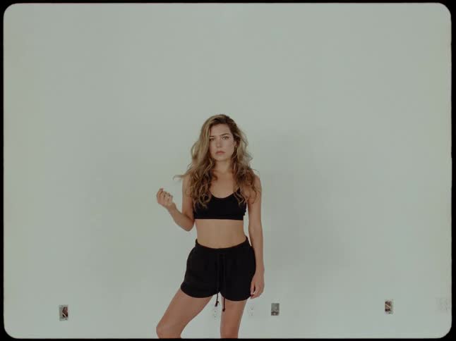 A woman in black athletic wear stands still, looking at the camera against a plain white wall in a retro-style video