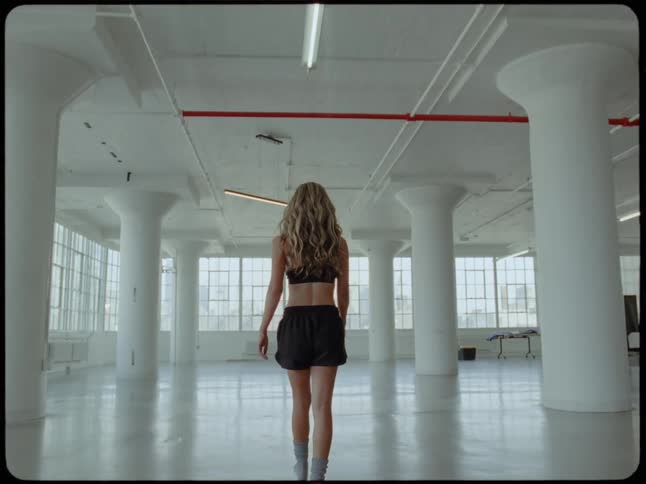 A woman in athletic wear walks away from the camera in slow motion through a vast, empty industrial loft