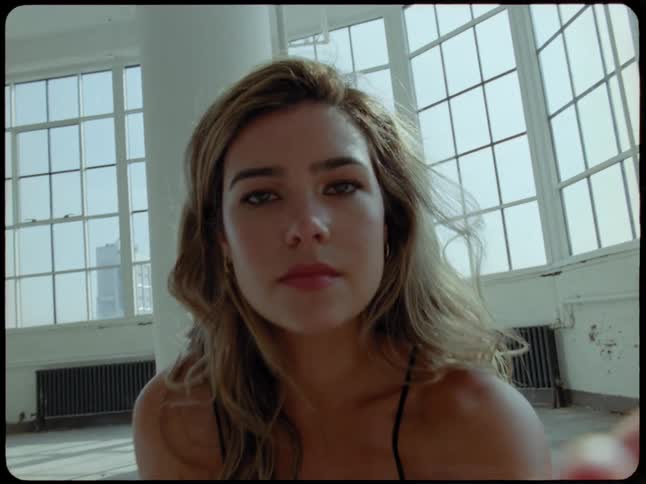 An expressive woman looks directly into the camera, reaches her hand towards the lens, and begins to dance in a sunlit urban loft
