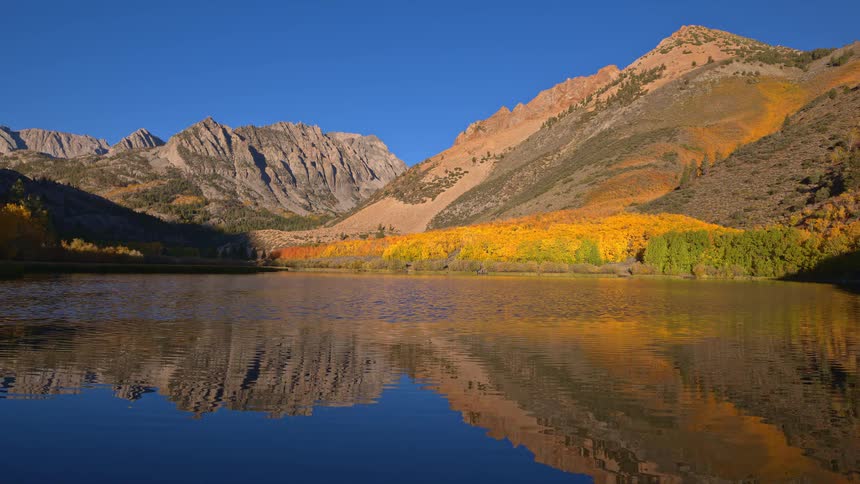 A serene mountain lake perfectly reflects the golden autumn foliage and rugged peaks under a clear blue sky