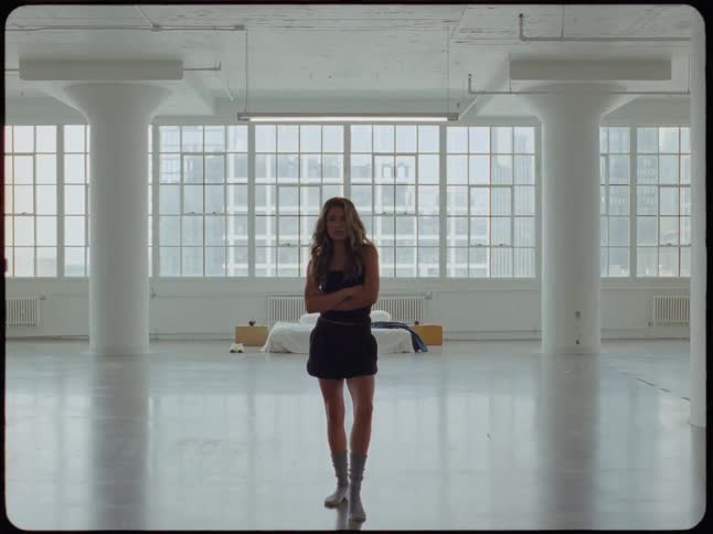 A confident woman stands with her arms crossed in the center of a vast, empty industrial loft apartment with large windows overlooking the city