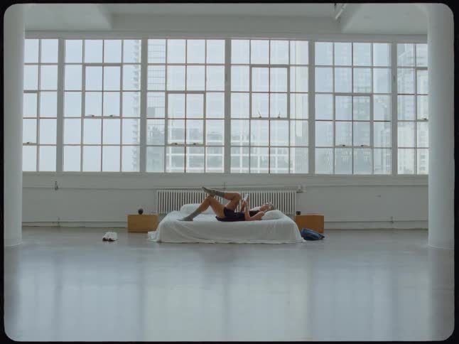 A woman reads a book while relaxing on her bed in a spacious, sunlit minimalist city loft apartment