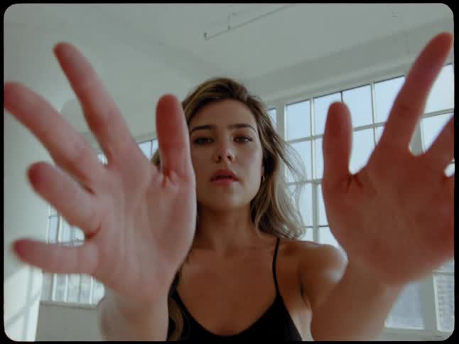 An intense young woman in workout clothes walks towards the camera and reaches for it in a sunlit city loft