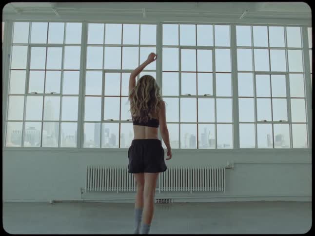 A young woman in athletic wear walks in slow motion through a bright, empty loft and looks out the large window at the city skyline