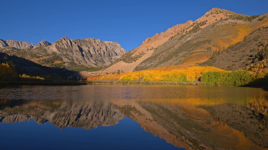 Majestic mountains and vibrant yellow autumn foliage are perfectly reflected in the still, blue water of a tranquil alpine lake