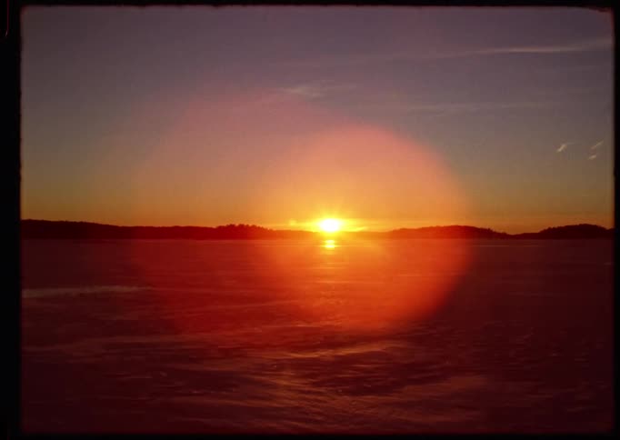 Vintage-style footage captures a serene sunset with a prominent lens flare over a vast, frozen winter landscape