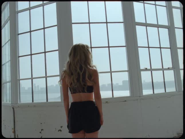 A young woman in a sunlit loft walks to a large window overlooking the city, then turns and spins in slow motion