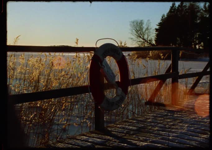 Vintage film footage pans from a bright sun flare across a frozen, snowy lake to a life preserver on a wooden dock at sunset