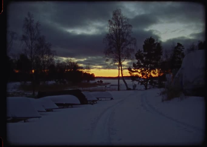 A vintage Super 8 film captures a tranquil winter sunset over a snowy landscape with covered boats by the water