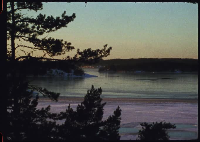 Vintage Super 8 film captures a tranquil winter landscape with an icy lake reflecting the sunset sky behind silhouetted trees