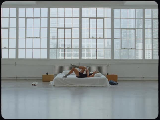 A woman reads a book while lying on a bed in the center of a spacious, minimalist white loft apartment with a large wall of windows