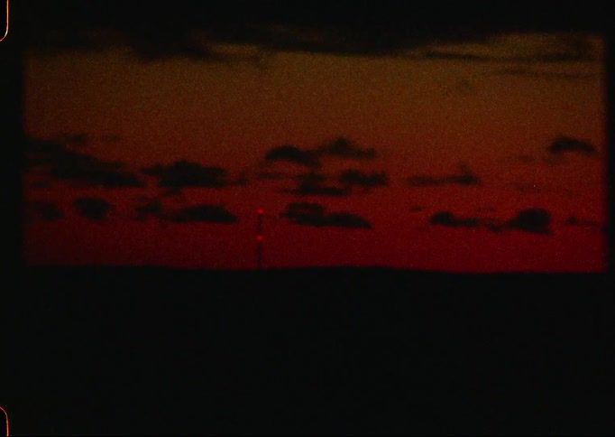 A blinking red light on a distant tower punctuates a deep red sunset in this grainy, moody vintage film shot