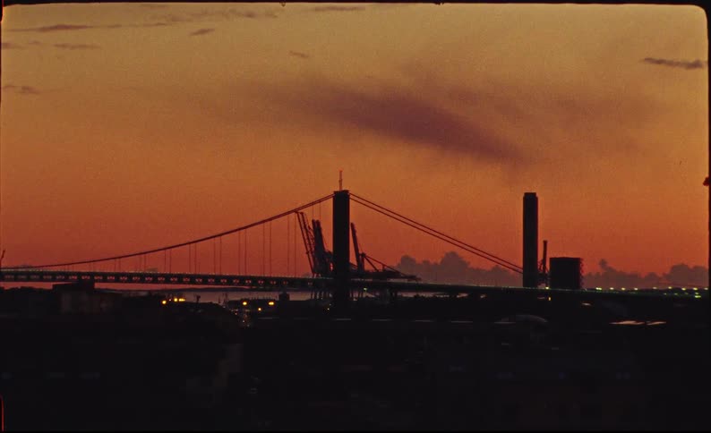 An industrial cityscape and bridge are silhouetted against a dramatic orange sky at sunset in this vintage film footage
