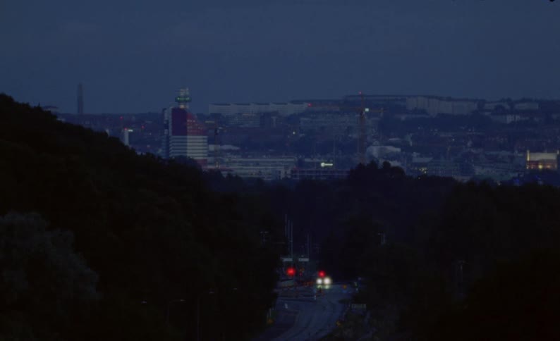 An early morning tram with bright headlights approaches through a dark, wooded area with the city skyline of Gothenburg visible in the background at dawn