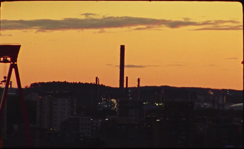 Vintage film captures the silhouette of an industrial city with a prominent smokestack against a warm, orange sunset sky