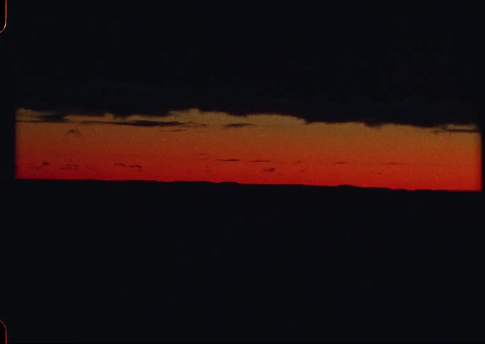 A sliver of a dramatic red and orange sunset sky is seen between dark clouds and a silhouetted landscape on grainy vintage film
