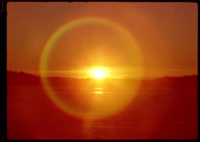 A time-lapse of a glowing sunrise over a calm lake, with a beautiful rainbow lens flare appearing around the sun on vintage film