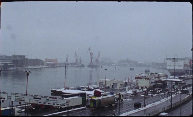 Vintage film captures a snowy, overcast day at an industrial harbor with traffic moving along the docks