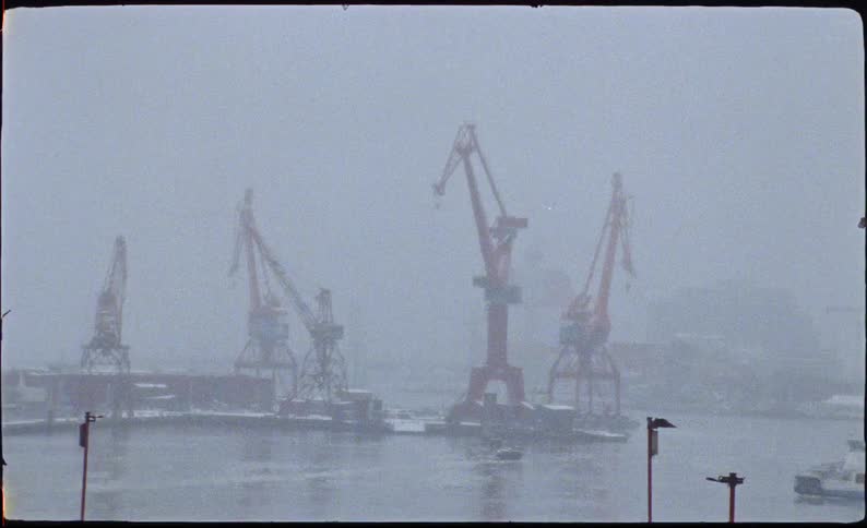 Industrial cranes stand idle in a foggy harbor during a heavy snowstorm on a winter day