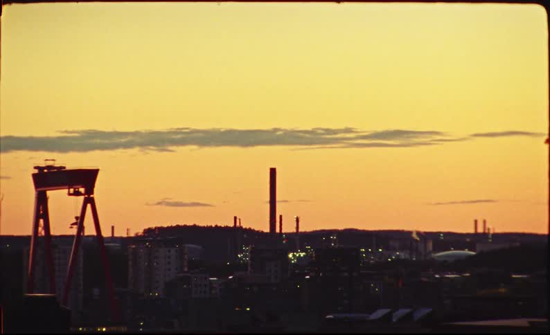 A vintage film shot captures an industrial city skyline with a large crane and smokestacks silhouetted against a warm golden sunset