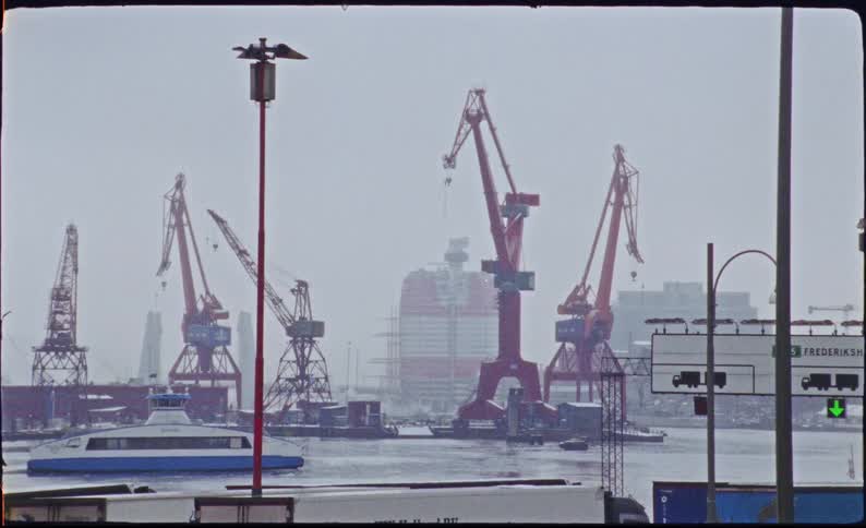 A vintage film shot of a ferry sailing past large red cranes in an industrial harbor on a gray, overcast day