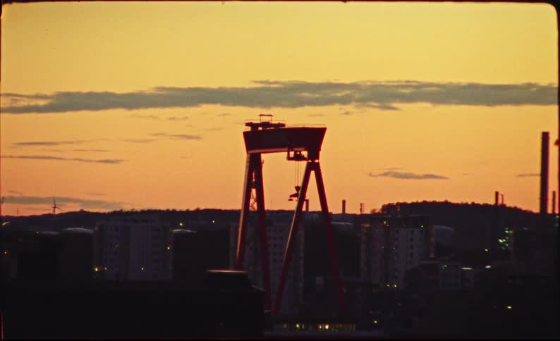 A large industrial shipyard crane stands silhouetted against a vibrant orange sunset sky over a city
