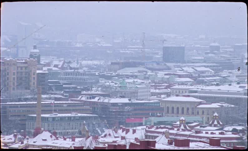 Snow falls gently over the rooftops of a European city on a cold and hazy winter day, captured on vintage film