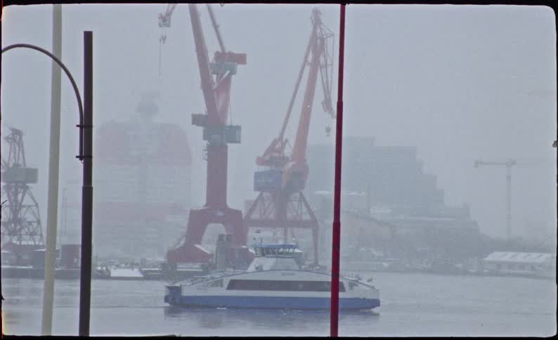 A 16mm analog film shot shows a ferry boat crossing an industrial harbor with large red cranes on a snowy and foggy winter day