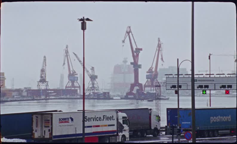 Large shipping cranes tower over a city harbor on a cold and snowy winter day as cargo trucks wait nearby