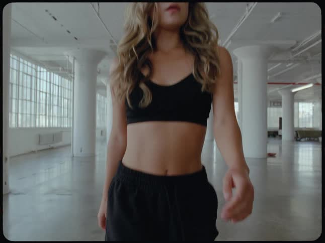 A fit young woman in black athletic wear walks confidently towards the camera in slow motion inside a bright, empty industrial loft