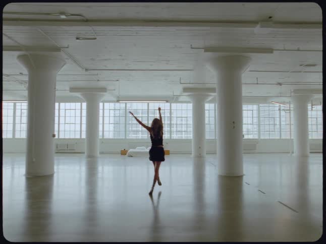 A young woman walks and dances with expressive movements through a vast, empty white loft space